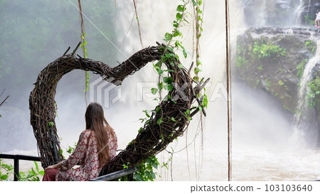 a young woman on a swing in the shape of a heart, enjoying the rainforest and looking at the high powerful waterfall Tegenungan. Rear view of a tourist girl on the Indonesian island of Bali a young woman on a swing in the shape of a heart, enjoying the rainforest and looking at the high powerful waterfall Tegenungan. Rear view of a tourist girl on the Indonesian island of Bali 103103640