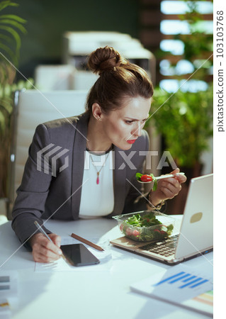 business woman in green office eating salad and using phone 103103768