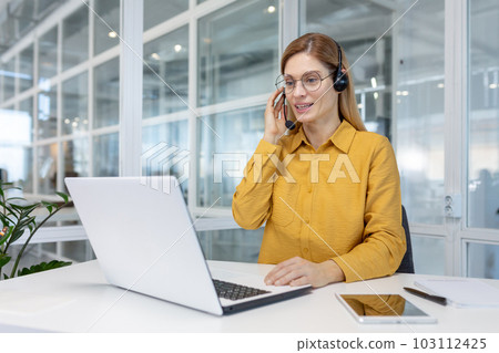 Portrait of a young woman in a headset working in an office center online from a laptop. Portrait of a young woman in a headset working in an office center online from a laptop. 103112425