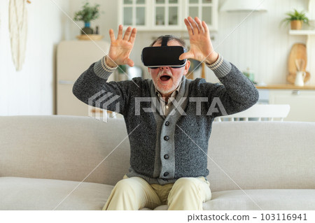 Excited mature senior man wearing using virtual reality metaverse VR glasses headset at home. Grandfather touching air during VR experience on virtual reality helmet. Simulation hi-tech videogame 103116941