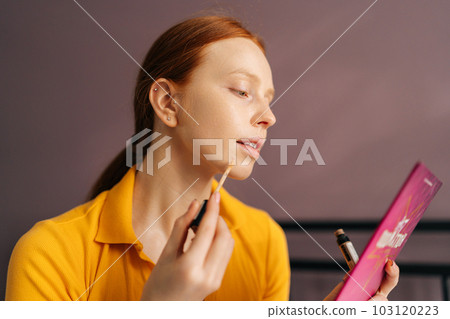 Closeup side view of attractive young woman applying foundation on face to correct skin and hide pimples near lips in bedroom. Red-hared female doing makeup putting base tone cream using concealer. Closeup side view of attractive young woman applying foundation on face to correct skin and hide pimples near lips in bedroom. Red-hared female doing makeup putting base tone cream using concealer. 103120223