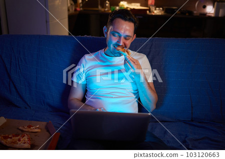 Handsome young man eating unhealthy food while watching movie at night. Handsome young man eating unhealthy food while watching movie at night. 103120663