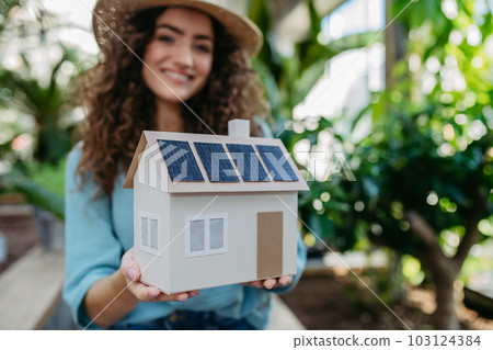Young woman in botanical garden holding model of house with solar panels, concept of green energy. Young woman in botanical garden holding model of house with solar panels, concept of green energy. 103124384