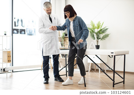 Specialist helping woman in leaning on cane in exam room 103124601