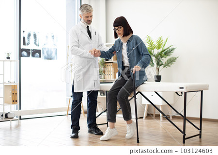 Specialist helping woman in leaning on cane in exam room Specialist helping woman in leaning on cane in exam room 103124603