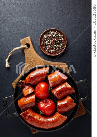 Fried pork sausage cut into pieces in a cast iron pan with tomatoes and peppercorns on a wooden cutting board 103125580