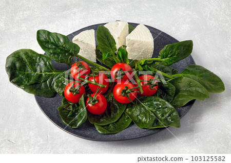 Cherry tomatoes on a branch with spinach and mozzarella cheese on a ceramic gray plate on a light stone table Cherry tomatoes on a branch with spinach and mozzarella cheese on a ceramic gray plate on a light stone table 103125582