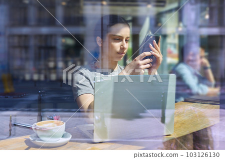 Young female inside cafe with cup of coffee by laptop smartphone looking through window Young female inside cafe with cup of coffee by laptop smartphone looking through window 103126130