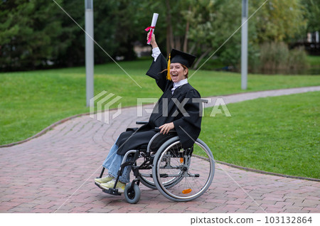 Caucasian woman in a wheelchair in a graduate costume rejoices at receiving a diploma. Caucasian woman in a wheelchair in a graduate costume rejoices at receiving a diploma. 103132864