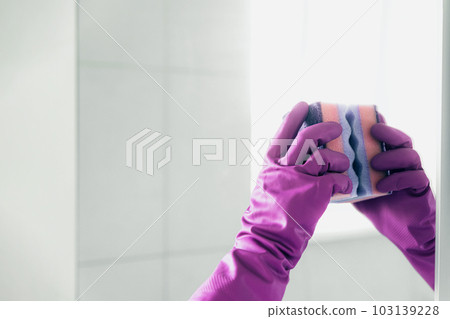 Cropped view of housekeeper woman hand in pink rubber glove polishing, cleaning, swiping mirror with cloth rag, spraying cleaner liquid while cleaning bathroom at home. Housekeeping, cleaning service 103139228
