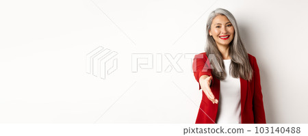 Professional asian businesswoman with grey hair, saying hello, stretch out hand for handshake and smiling, standing over white background 103140488