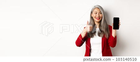 Satisfied asian elderly businesswoman showing blank smartphone screen and thumb-up, praising online promotion or company app, standing over white background 103140510