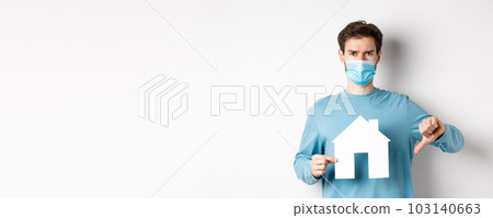Covid and real estate concept. Disappointed young man in medical mask showing paper house cutout and thumb down, dislike broker agency, standing displeased over white background Covid and real estate concept. Disappointed young man in medical mask showing paper house cutout and thumb down, dislike broker agency, standing displeased over white background 103140663