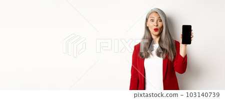 Amused senior woman in red blazer, looking fascinated and showing blank smartphone screen, standing over white background 103140739