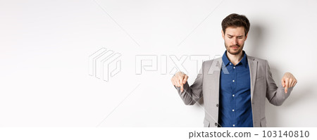 Unimpressed young businessman frowning disappointed, pointing and looking down at bad promotion, standing displeased on white background Unimpressed young businessman frowning disappointed, pointing and looking down at bad promotion, standing displeased on white background 103140810