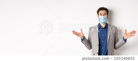 Health, pandemic and business concept. Confused businessman in medical mask and suit spread hands sideways and look puzzled, cant understand, white background 103140855