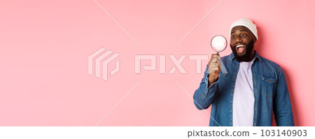 Happy african-american man looking through magnifying glass, smiling amazed, standing against pink background 103140903