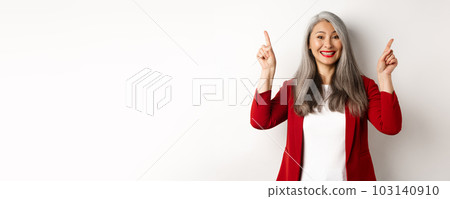 Happy mature woman in red blazer and makeup, smiling and showing advertisement on top, pointing fingers up at logo, white background Happy mature woman in red blazer and makeup, smiling and showing advertisement on top, pointing fingers up at logo, white background 103140910