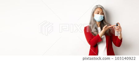 Covid, pandemic and business concept. Businesswoman in red blazer and face mask using hand sanitizer to clean and disinfect, standing over white background Covid, pandemic and business concept. Businesswoman in red blazer and face mask using hand sanitizer to clean and disinfect, standing over white background 103140922