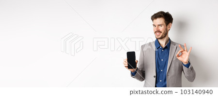 E-commerce and online shopping concept. Smiling businessman show okay sign and empty smartphone screen, demonstrate his account, standing on white background 103140940