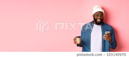 Image of stylish Black man hipster drinking takeaway coffee, reading message on phone and smiling, standing over pink background 103140970
