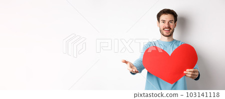Handsome man in love making confession to you, pointing hand at camera, holding big red heart cutout on valentines day, singing romantic serenade, standing over white background 103141118