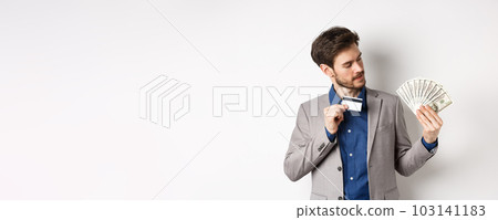 Successful man showing plastic credit card and looking at dollar bills fan, making money deposit, standing on white background 103141183