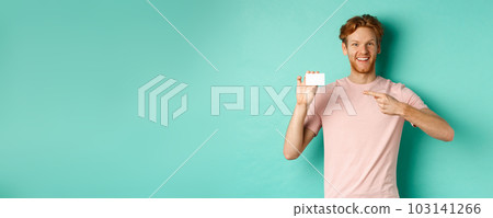 Attractive adult man with beard and red hair pointing finger at plastic credit card, smiling pleased at camera, standing over turquoise background 103141266