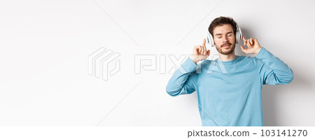 Relaxed young man enjoying favorite song, listening music in headphones with closed eyes and calm face, standing over white background 103141270