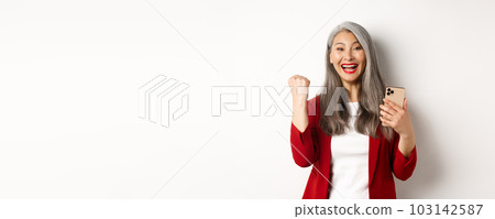 Asian old woman winning online, holding smartphone and making fist pump gesture to celebrate win, triumphing and smiling, standing over white background 103142587