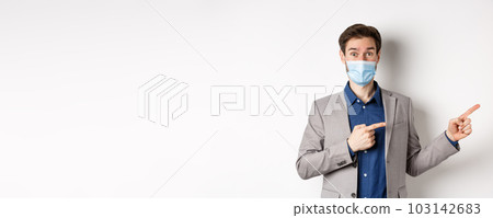 Covid-19, pandemic and business concept. Excited male entrepreneur in medical mask and suit pointing fingers right and looking curious, white background 103142683