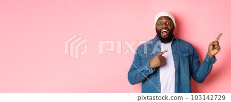 Excited and amazed Black man showing awesome offer, pointing fingers right at copy space, standing in hipster beanie and denim shirt on pink background Excited and amazed Black man showing awesome offer, pointing fingers right at copy space, standing in hipster beanie and denim shirt on pink background 103142729