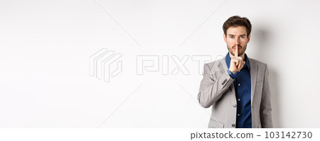 Handsome macho man in suit shushing at camera, press finger to lips, ask keep voice down, tell secret, standing against white background 103142730