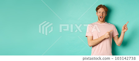 Excited guy in pink t-shirt showing advertisement on mint background, looking amazed at camera and pointing at upper right corner 103142738