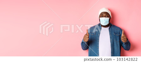 Covid-19, lifestyle and quarantine concept. Satisfied Black man in face mask showing thumbs-up, standing over pink background 103142822