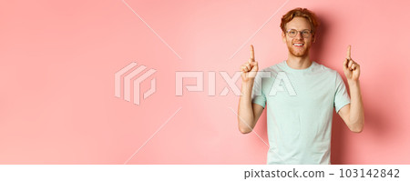 Attractive young man in glasses and t-shirt showing promo deal, pointing fingers up and smiling at camera, standing over pink background 103142842