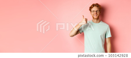 Cheerful redhead man in glasses and t-shirt, showing thumbs up with satisfied face, showing positive reaction, approve and agree with you, standing over pink background 103142928