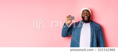 Shopping concept. Amazed Black guy showing credit card, staring at camera impressed, recommending bank, standing over pink background Shopping concept. Amazed Black guy showing credit card, staring at camera impressed, recommending bank, standing over pink background 103142966