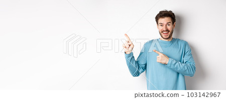 Happy caucasian man in casual blue sweatshirt, pointing fingers at upper left corner, showing link or logo on white background, smiling at camera 103142967