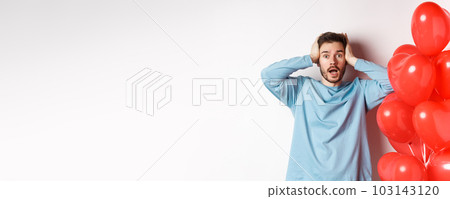 Confused boyfriend holding hands on head and panicking on Valentines holiday, alarmed with romantic presents on lovers day, standing near hearts balloon over white background 103143120