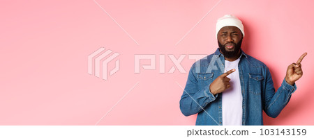 Disappointed and skeptical african-american guy complaining, cringe from disgust, frowning and grimacing from dissatisfaction, pointing fingers right, pink background 103143159