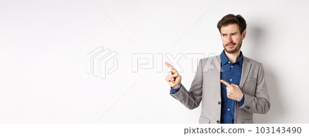 Displeased businessman in suit pointing and looking left with skeptical smirk, dislike banner, disapprove promo offer, standing on white background Displeased businessman in suit pointing and looking left with skeptical smirk, dislike banner, disapprove promo offer, standing on white background 103143490