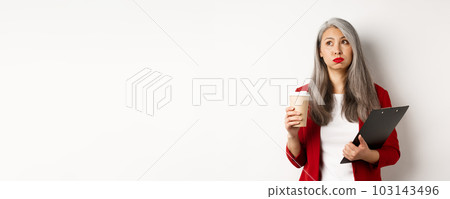 Tired asian female office worker holding clipboard and paper cup, drinking coffee and exhaling with exhausted face, standing over white background 103143496