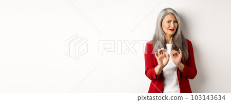 Disgusted asian businesswoman with grey hair, wearing red blazer and makeup, rejecting something disgusting, showing stop sign, white background 103143634