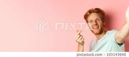 Handsome young man with beard and red hair taking selfie against pink background, stretch out hand to hold smartphone camera and showing korean heart gesture, pink background 103143648