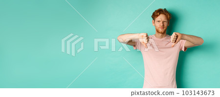 Disappointed guy with red hair showing thumbs-down, frowning and looking skeptical, epxress dislike and disapproval, standing over turquoise background 103143673