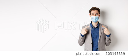 Covid-19, pandemic and business concept. Excited businessman in suit pointing at his medical mask and smiling with eyes, white background 103143839