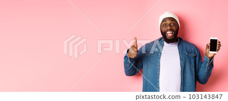 Online shopping and technology concept. Happy african-american hipster man showing thumbs-up and mobile screen, praising app, standing over pink background Online shopping and technology concept. Happy african-american hipster man showing thumbs-up and mobile screen, praising app, standing over pink background 103143847