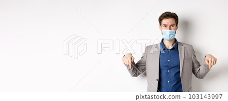 Covid-19, pandemic and business concept. Handsome businessman in medical mask and suit, pointing fingers down and showing company logo, white background 103143997