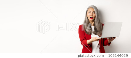 Business. Impressed asian old woman working on laptop, staring at camera surprised, standing over white background Business. Impressed asian old woman working on laptop, staring at camera surprised, standing over white background 103144029